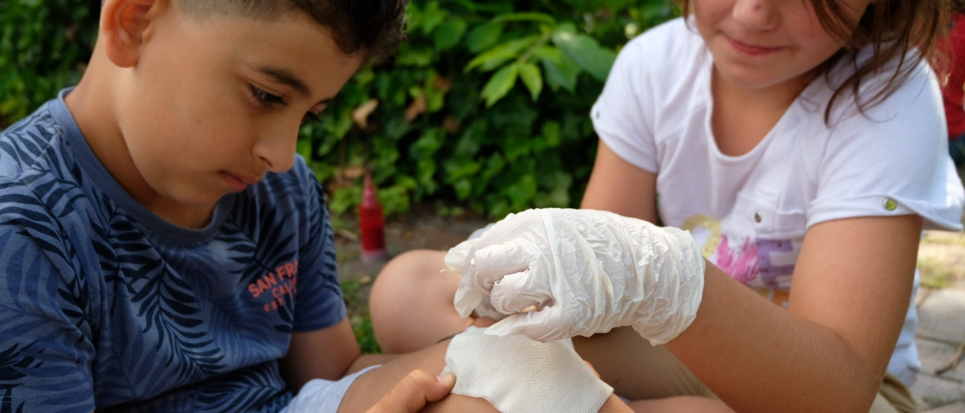 Ein Kind mit verletztem Knie wird von einem anderen Kind verarztet.