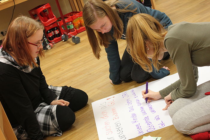 Drei Mädchen knien im Kreis um ein Plakat. Das Plakat ist in bunten Buchstaben gestaltet und dafür gedacht, neue Gruppenleitungen zu begeistern.