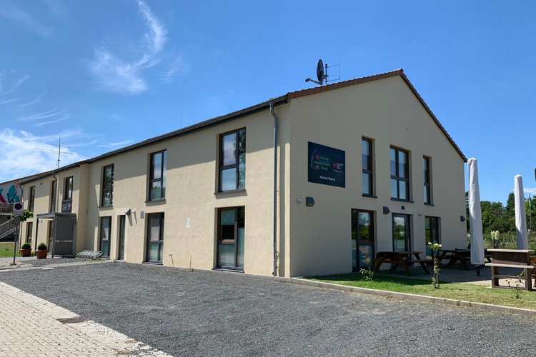jrk-nds_haus-des-jugendrotkreuzes_stadthaus.jpg Zu sehen ist das Stadthaus: Ein langgezogenes Gebäude mit zwei Etagen und einer Terrasse mit Bänken und Sonnenschirmen.