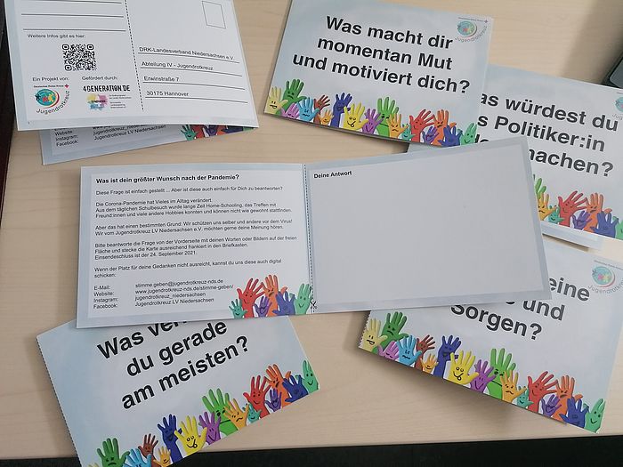 Sechs Klapppostkarten liegen auf einem Tisch. Eine ist aufgeklappt, von anderen ist die Vorderseite zu sehen. Darauf abgebildet sind bunte Hände mit jeweils unterschiedlichen, aufgemalten Gesichtsausdrücken und dazu unterschiedliche Fragen.